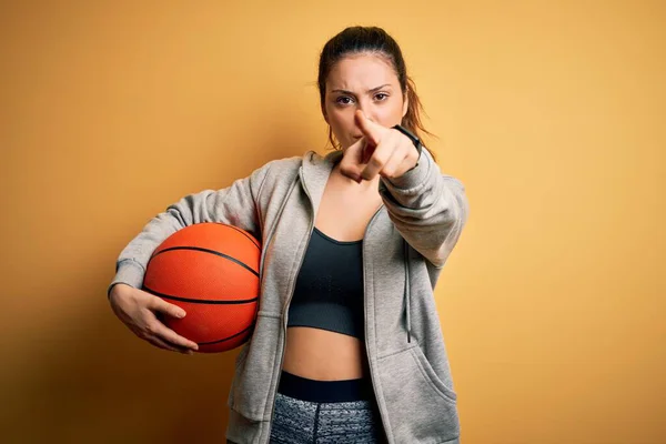 年轻美丽的黑发女运动员 背景是黄色的 手握篮球 手指指向摄像机和你 正面是积极自信的手势 — 图库照片