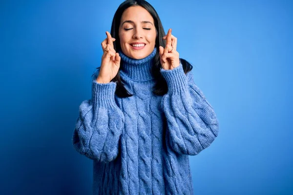 Young brunette woman with blue eyes wearing casual turtleneck sweater gesturing finger crossed smiling with hope and eyes closed. Luck and superstitious concept.