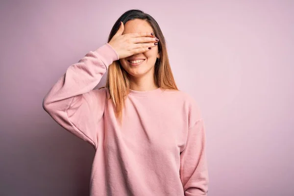Beautiful blonde woman with blue eyes wearing sweater and glasses over pink background smiling and laughing with hand on face covering eyes for surprise. Blind concept.