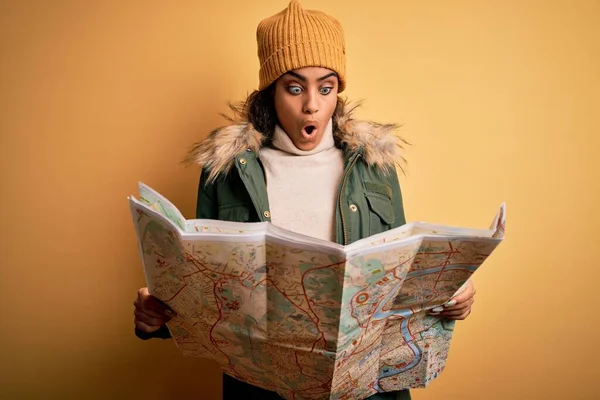 African american skier tourist girl wearing snow sportswear and ski goggles holding city map scared in shock with a surprise face, afraid and excited with fear expression