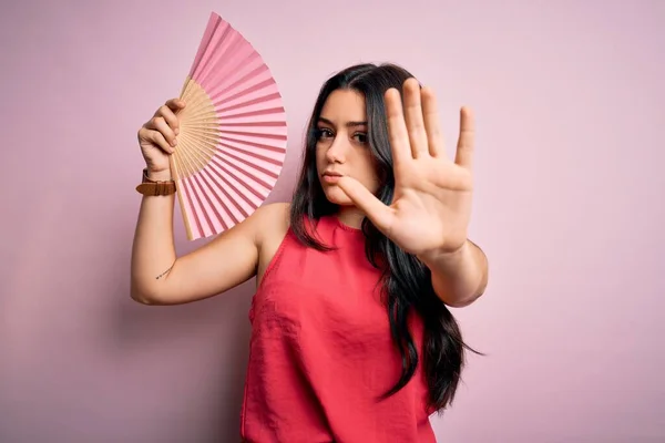 Young brunette woman holding hand fan for fresh air over pink isolated background with open hand doing stop sign with serious and confident expression, defense gesture