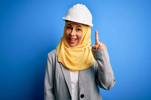 Middle age brunette architect woman wearing muslim traditional hijab and security helmet pointing finger up with successful idea. Exited and happy. Number one.