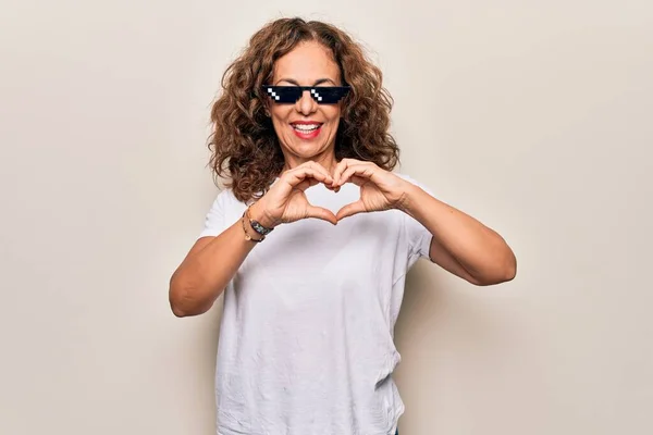 Middle age beautiful woman wearing funny thug life sunglasses over white background smiling in love doing heart symbol shape with hands. Romantic concept.