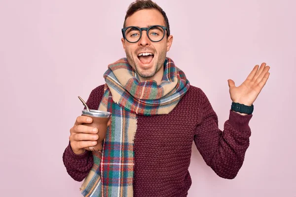 Young handsome man with blue eyes drinking cup of hot mate tea over pink background very happy and excited, winner expression celebrating victory screaming with big smile and raised hands