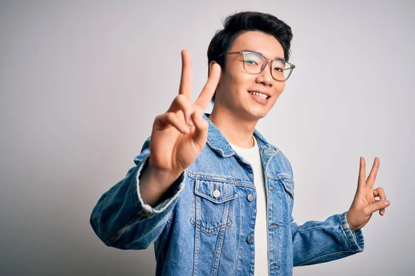 Young handsome chinese man wearing denim jacket and glasses over white background smiling looking to the camera showing fingers doing victory sign. Number two.