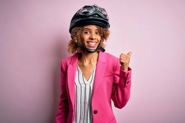アフリカ系アメリカ人オートバイ女性とともに巻き毛を身に着けています本ヘルメット上のピンクの背景笑顔で幸せな顔を見てと親指アップで側面を指して — ストック写真