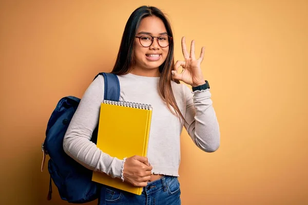 Young smart asian student girl wearing backpack holding notebook over yellow background doing ok sign with fingers, excellent symbol