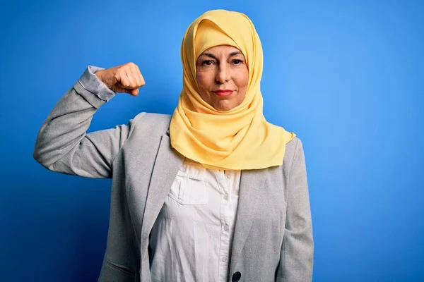 Middle age brunette business woman wearing muslim traditional hijab over blue background Strong person showing arm muscle, confident and proud of power