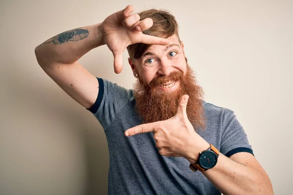 Handsome Irish redhead man with beard and arm tattoo standing over isolated background smiling making frame with hands and fingers with happy face. Creativity and photography concept.