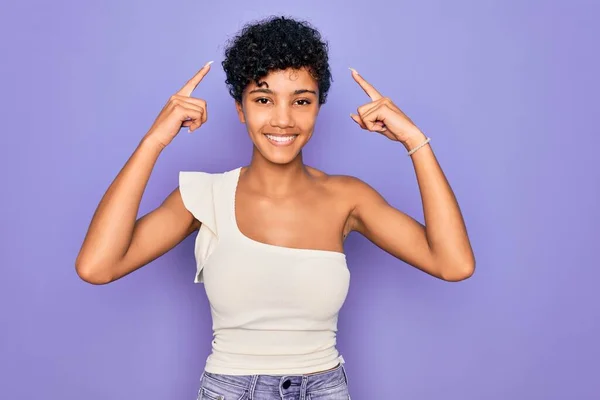 年轻美丽的非洲裔美国女人 身穿浅紫色背景的休闲T恤 用双手指指着头微笑 很好的主意或想法 好的记忆力 — 图库照片