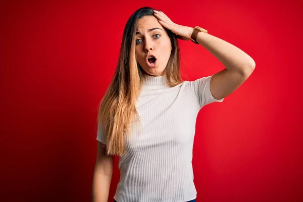 Beautiful blonde woman with blue eyes wearing casual white t-shirt over red background surprised with hand on head for mistake, remember error. Forgot, bad memory concept.