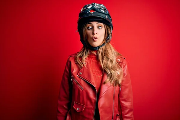赤い背景の上に元ヘルメットを身に着けている若い美しいブルネットのオートバイの女性は 狂気とコミカルなジェスチャーで魚の顔を作る 面白い表現 — ストック写真