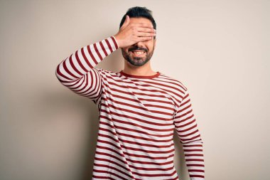 Sakallı, sıradan çizgili tişörtler giyen, beyaz arka planda duran, gülümseyen ve yüzünde şaşkınlık için gözlerini kapatan genç yakışıklı bir adam. Kör kavram.