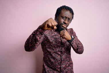 Genç ve yakışıklı Afro-Amerikalı bir adam, günlük gömleğini giymiş pembe arka planda duruyor yumruklarını yumrukluyor, saldırıyor, saldırıyor, tehdit ediyor ve şiddet uyguluyor.