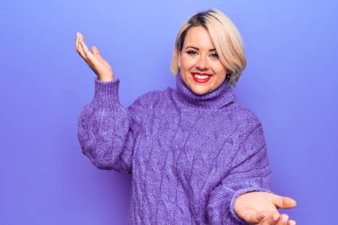 Güzel sarışın ve cüsseli bir kadın mor arka plan üzerine balıkçı yaka kazak giyip gülümsüyor ve kollarını açıp gülümsüyor. Hoş geldin, pozitif ve kendinden emin selamlar.