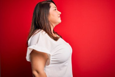 Güzel esmer ve cüsseli bir kadın izole edilmiş kırmızı arka plan üzerine gündelik tişört giyer, rahat bir görünüm takınır, kendine güvenen bir gülümsemeyle doğal bir yüzle poz verir..