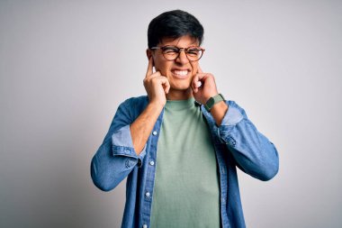 Gürültülü müziğin gürültüsü için günlük gömlek ve gözlük takan genç yakışıklı bir adam izole edilmiş beyaz arka planda kulakları parmaklarıyla kapatıyor. Sağır kavramı.