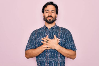 Pembe arka planda hippi tarzı giyinmiş genç, yakışıklı bohem bir adam elleri göğsünde, gözleri kapalı ve yüzünde minnet dolu bir jest ile gülümsüyor. Sağlık kavramı.