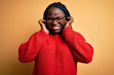 Sarı arka plan üzerine gündelik süveter giyen Afrikalı Amerikalı artı cüsseli kadın yüksek sesli müzik sesi için rahatsız edici ifadeleri olan parmaklarıyla kulaklarını kapatıyor. Sağır kavramı.
