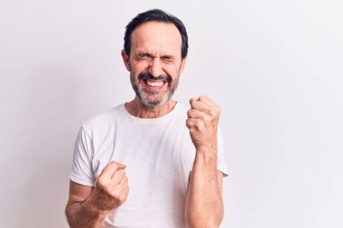 Orta yaşlı yakışıklı bir adam, günlük tişört giyip izole edilmiş beyaz arka planda dikilmiş şaşırmış ve şaşırmış bir şekilde kolları havada ve gözleri kapalı bir şekilde başarıyı kutluyor.