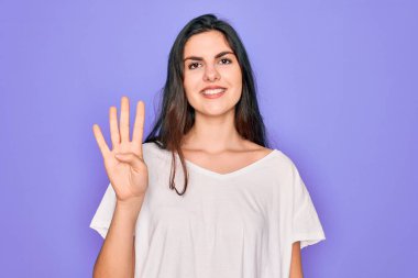 Genç, güzel esmer kadın, mor arka plan üzerine gündelik beyaz tişört giyip parmaklarıyla dört numarayı gösterirken kendinden emin ve mutlu bir şekilde gülümsüyor..