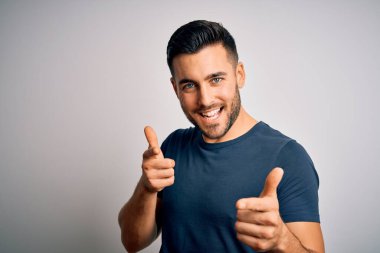 İzole edilmiş beyaz arka plan üzerinde dikilmiş tişört giyen genç yakışıklı adam mutlu ve komik yüzlü parmaklarını kameraya doğrultuyor. İyi enerji ve titreşimler.