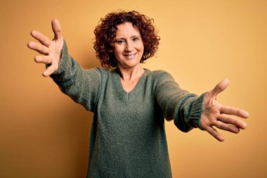 Orta yaşlı güzel kıvırcık saçlı kadın izole edilmiş sarı arka planda sıradan bir süveter giyiyor ve kameraya sarılıp gülümsüyor. Mutluluğu kucaklayan neşeli bir ifade..