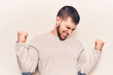 Genç yakışıklı bir adam gündelik süveter giyiyor. Çok mutlu ve heyecanlı. Kolları havada, gülümseyen ve başarı için bağıran kazanan bir jest yapıyor. kutlama konsepti. 