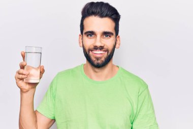 Sakallı, bir bardak su içen genç yakışıklı adam pozitif ve mutlu görünüyor ve kendine güvenen bir gülümsemeyle dişlerini gösteriyor. 