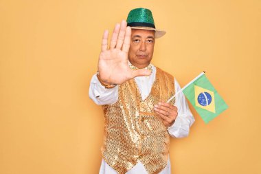 Orta yaşlı, kır saçlı, Brezilya karnavalı kıyafeti giyen, elinde Brezilya bayrağı tutan, ciddi ve kendinden emin bir ifadeyle dur işareti yapan, savunma hareketi yapan yaşlı bir adam.