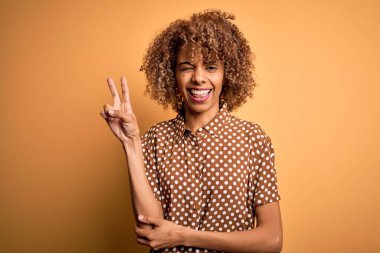 Genç, güzel, Afro-Amerikan bir kadın, günlük gömleğini giymiş, sarı arka planda dikiliyor, gülümsüyor ve kameraya bakıp zafer işareti yapıyor. İki numara..
