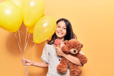 Sevimli İspanyol çocuk mutlu gülümsüyor. Yüzünde gülücüklerle balon tutarken ve ayıcık dondurma yerken izole edilmiş sarı arka plan