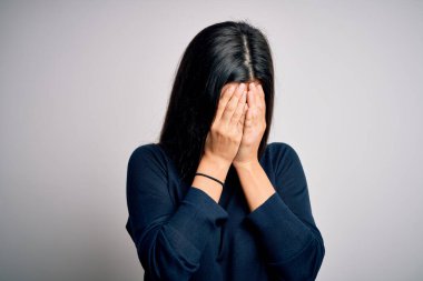 Genç, güzel, esmer bir kadın beyaz arka planda dikilip ağlarken suratını ellerle kapatan sıradan bir kazak giyiyor. Depresyon kavramı.