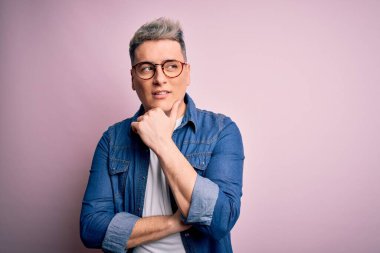 Pembe izole edilmiş arka plan üzerinde gözlük ve kot ceket giyen yakışıklı, soru soran, düşünceli bir genç adam. Düşünceli bir yüzle gülümsüyor. Şüphe kavramı.