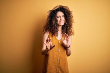 Güzel esmer kadın tatilde. Kıvırcık saçlı ve piercing takıyor. Şapka ve elbise giyiyor. Eller havada