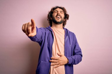 Genç, yakışıklı, sakallı, pembe arka planda sıradan bir kazak giyen, sana gülen, eli vücudunun üzerinde kamerayı işaret eden, utanç ifadesi olan yakışıklı bir adam.