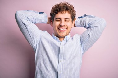 Sarışın, kıvırcık saçlı, beyaz arka planda çizgili gömlek giyen, kolları ve elleri başının arkasında, mutlu bir şekilde gülümseyen genç bir adam.