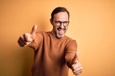 Orta yaşlı, beyaz, kahverengi süveter ve gözlük takan, sarı arka planda, olumlu bir el hareketi onaylayan, gülümseyen ve başarı için mutlu bir adam. Kazanan jest.