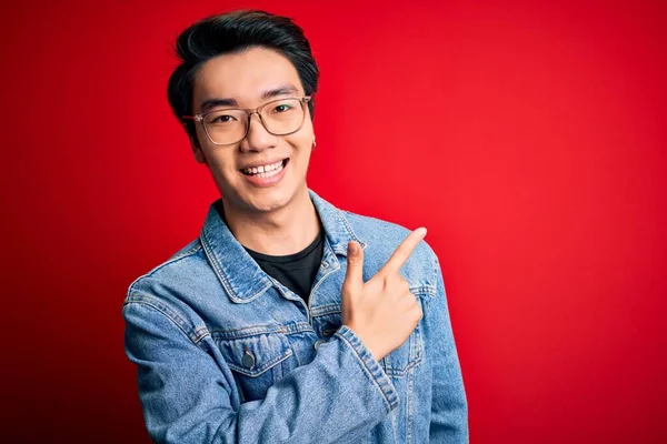 Young handsome chinese man wearing denim jacket and glasses over red background cheerful with a smile of face pointing with hand and finger up to the side with happy and natural expression on face