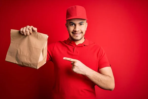Kırmızı Arka Planda Paket Yiyecek Taşıyan Genç Yakışıklı Teslimatçı Parmağıyla — Stok fotoğraf
