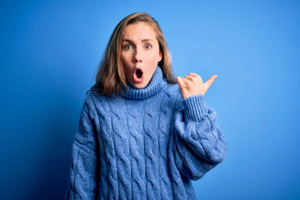 Young beautiful blonde woman wearing casual turtleneck sweater over blue background Surprised pointing with hand finger to the side, open mouth amazed expression.