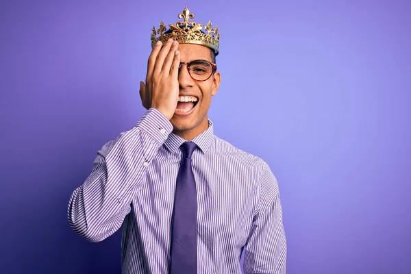 若いですハンサムなアフリカ系アメリカ人男性身に着けています黄金の冠の王以上紫色の背景カバー1目で手 顔に自信を持って笑顔と驚きの感情 — ストック写真