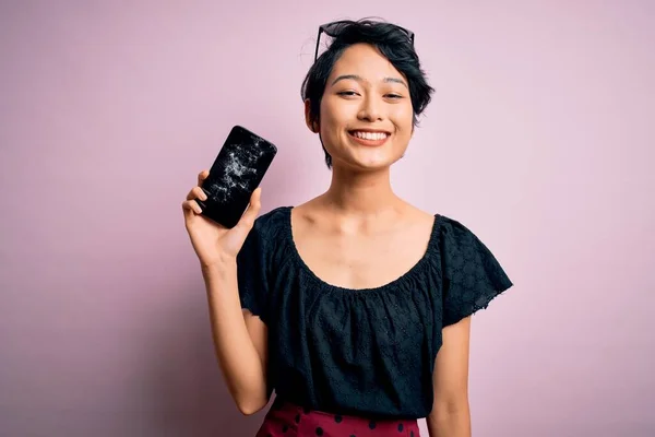 Young beautiful chinese woman holding broken smartphone showing craked screen with a happy face standing and smiling with a confident smile showing teeth