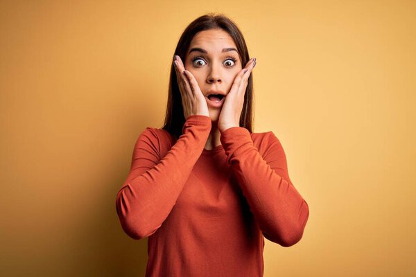 Young beautiful brunette woman wearing casual t-shirt standing over yellow background afraid and shocked, surprise and amazed expression with hands on face