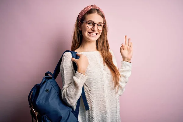 Young beautiful redhead student woman wearing backpack and glasses over pink background doing ok sign with fingers, excellent symbol