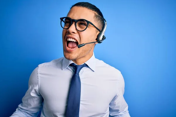 Young brazilian call center agent man wearing glasses and tie working using headset crazy and mad shouting and yelling with aggressive expression and arms raised. Frustration concept.