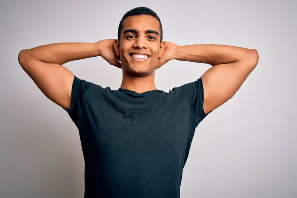 Young handsome african american man wearing casual t-shirt standing over white background relaxing and stretching, arms and hands behind head and neck smiling happy