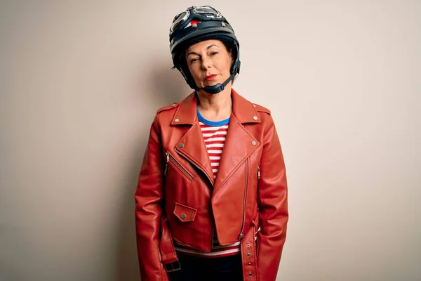 白い背景にバイク用ヘルメットとジャケットを身に着けている中年のオートバイ愛好家女性が顔に深刻な表情でリラックスしました シンプルで自然なカメラを見て — ストック写真