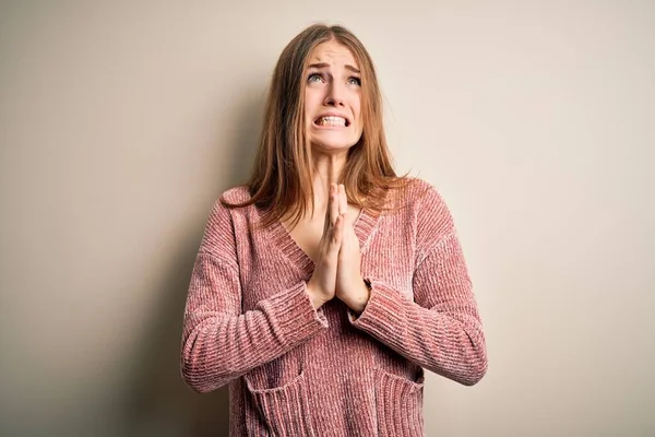 Young beautiful redhead woman wearing pink casual sweater over isolated white background begging and praying with hands together with hope expression on face very emotional and worried. Begging.