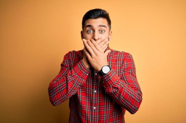 Izole sarı arka plan üzerinde duran rahat gömlek giyen genç yakışıklı adam hata için elleri ile ağız kapsayan şok. Gizli kavram.